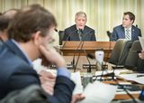 Steve Kelley sits at the testifiers' table and speaks to committee members whose faces are blurred in the foreground. Representative Long sits next to him and watches as he speaks.